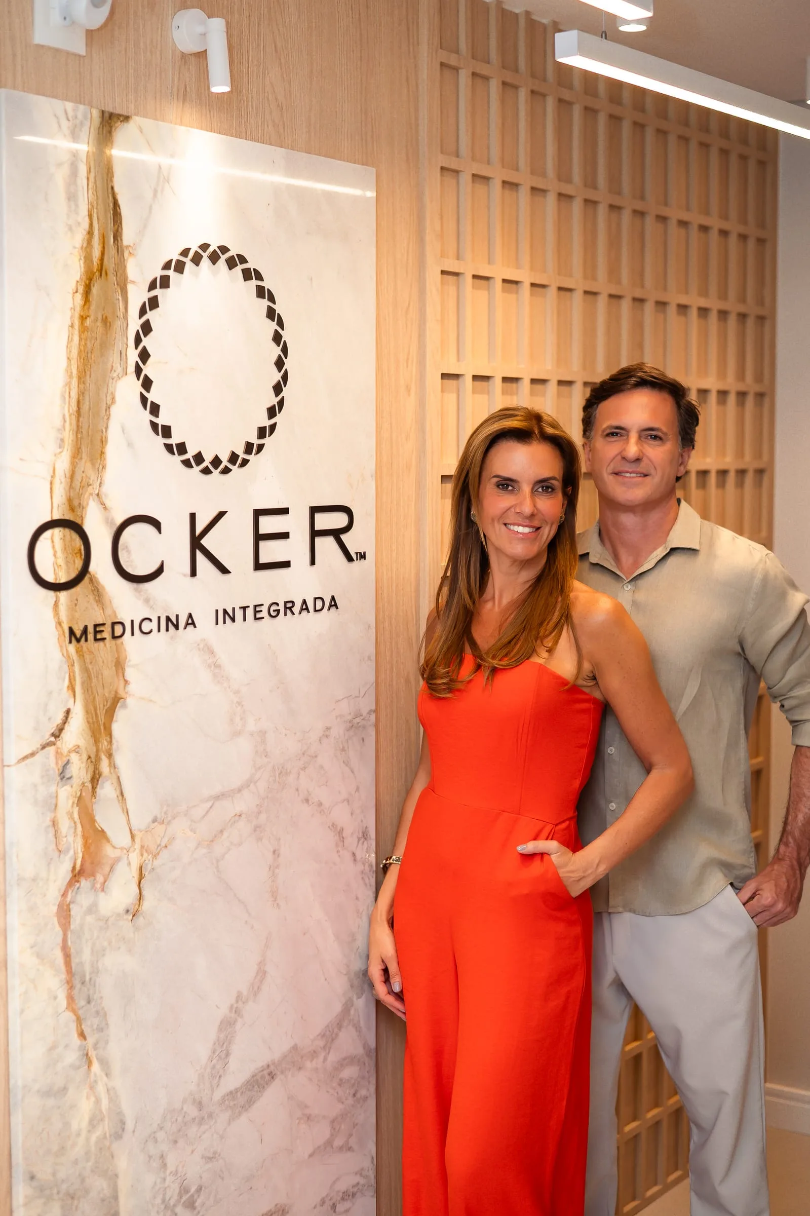 Dr. Guilherme e Dra. Juliana ao lado do painel com a logo da Ocker Medicina Integrada